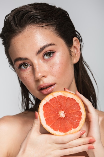 Retrato de belleza de una mujer joven sensual atractiva con el pelo largo morena mojado de pie aislado en gris, mostrando pomelo partido a la mitad, posando