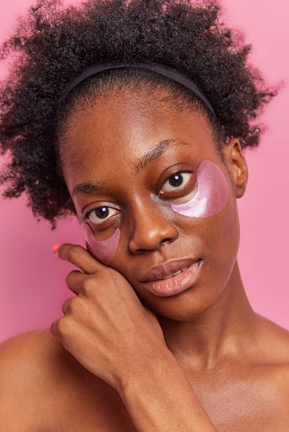 Retrato de belleza de mujer rizada de piel oscura toca suavemente la cara mira directamente a la cámara aplica parches hidratantes de hidrogel posa en el estudio con el cuerpo desnudo tiene poses de piel facial fresca en el interior.