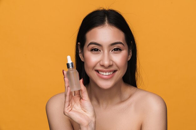 Retrato de belleza de mujer semidesnuda caucásica joven sonriendo y sosteniendo la botella de crema de suero facial aislada sobre pared amarilla