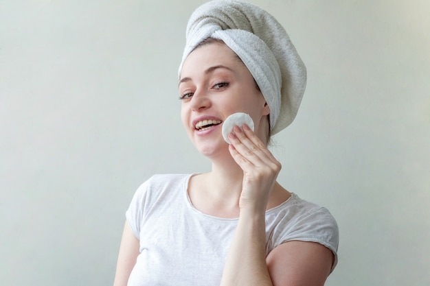 Retrato de belleza de mujer sonriente en toalla en la cabeza con piel suave y saludable quitando maquillaje con algodón aislado sobre fondo blanco.