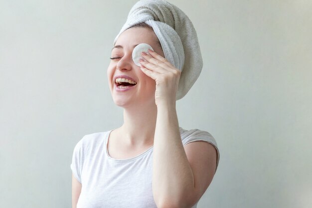 Retrato de belleza de mujer sonriente en toalla en la cabeza con piel suave y saludable quitando maquillaje con algodón aislado sobre fondo blanco.