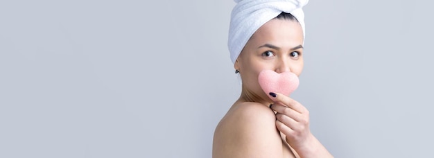 Retrato de belleza de mujer con toalla blanca en la cabeza con una esponja para un cuerpo a la vista de un corazón rosa Cuidado de la piel limpieza eco cosmética orgánica spa relax concepto