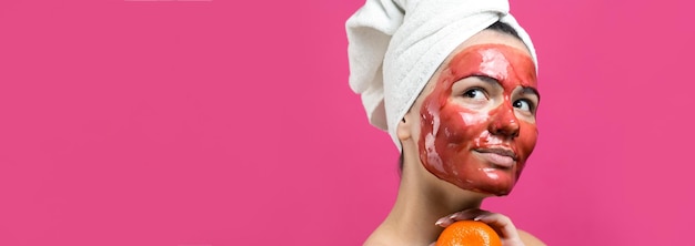 Retrato de belleza de mujer con toalla blanca en la cabeza con máscara nutritiva roja en la cara Cuidado de la piel limpieza eco cosmética orgánica spa relax concepto Una niña se para con la espalda sosteniendo una mandarina naranja