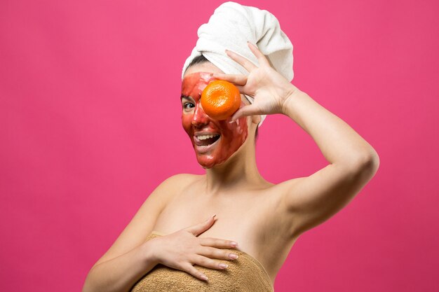 Retrato de belleza de mujer en toalla blanca en la cabeza con mascarilla nutritiva roja en la cara. Cuidado de la piel limpieza eco spa cosmético orgánico relajarse concepto. Una niña está de espaldas sosteniendo una mandarina naranja.