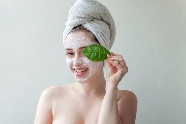 Retrato de belleza de mujer con toalla en la cabeza con máscara o crema nutritiva blanca en la cara y hoja verde en la mano, fondo blanco aislado.