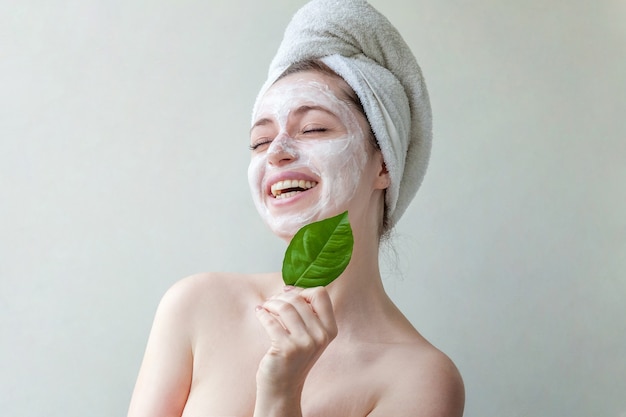 Retrato de belleza de mujer con toalla en la cabeza con máscara o crema nutritiva blanca en la cara y hoja verde en la mano, fondo blanco aislado.