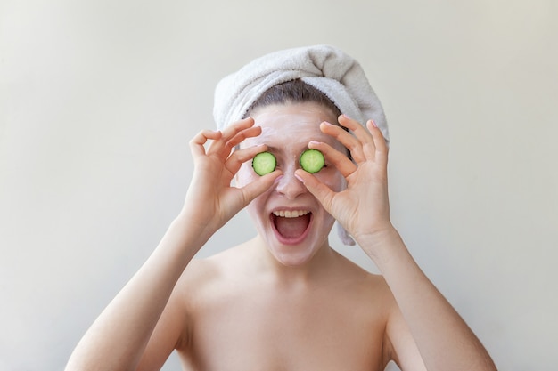 Foto retrato de belleza de mujer en toalla en la cabeza con mascarilla nutritiva blanca o crema en la cara con rodajas de pepino, fondo blanco aislado. cuidado de la piel limpieza eco cosmética orgánica spa relax concepto