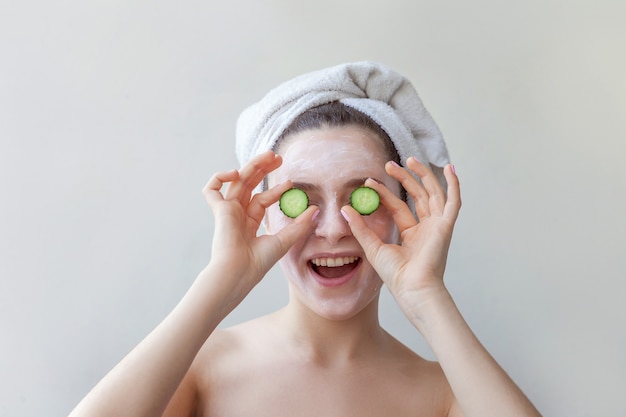 Retrato de belleza de mujer en toalla en la cabeza con mascarilla nutritiva blanca o crema en la cara con rodajas de pepino