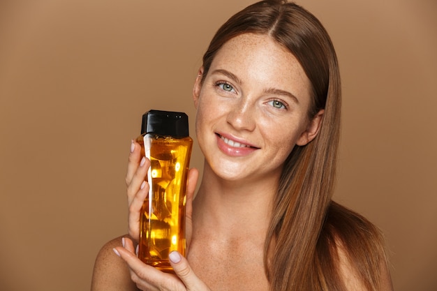 Retrato de la belleza de una mujer en topless joven sonriente con el pelo rojo largo que muestra una botella de champú aislada sobre la pared beige