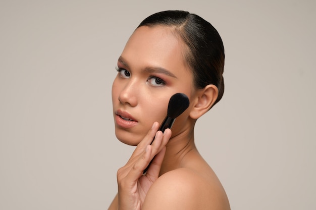 Retrato de la belleza de la pose de la mujer joven sensual atractiva asiática que sostiene el cepillo del colorete del maquillaje en su mejilla aislada sobre la pared gris