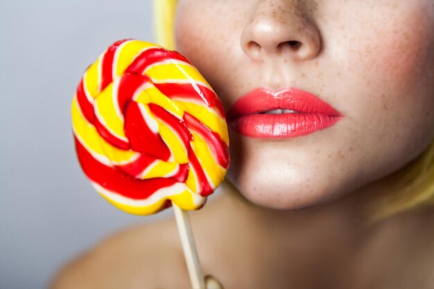 Retrato de la belleza del primer del modelo femenino joven lindo con las pecas, maquillaje rojo y peluca amarilla, sosteniendo el palillo del caramelo colorido cerca de sus labios rojos hermosos. tiro del estudio de interior, aislado en fondo gris.