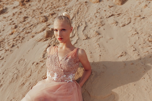 Retrato de belleza de primer plano de una hermosa joven rubia posando en vestido elegante en el desierto, acostado sobre arena dorada, mirando a cámara. Luz del atardecer.