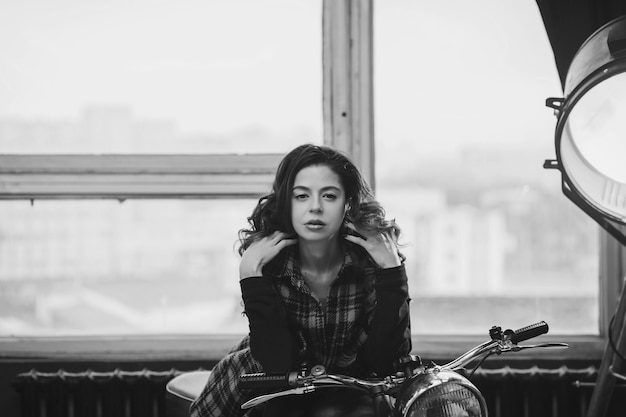 Retrato en blanco y negro de una chica de pelo rizado sexy con una camisa a cuadros cerca de una motocicleta en el garaje