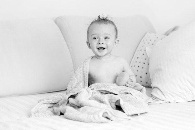 Retrato en blanco y negro de divertido bebé riendo sentado en la cama después de bañarse