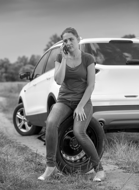 Retrato en blanco y negro de la joven conductora sentada junto al coche roto y hablando por teléfono