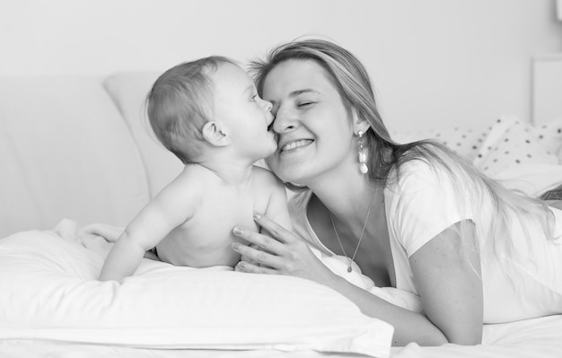 Retrato en blanco y negro de la madre alegre abrazando y besando a su bebé en la cama