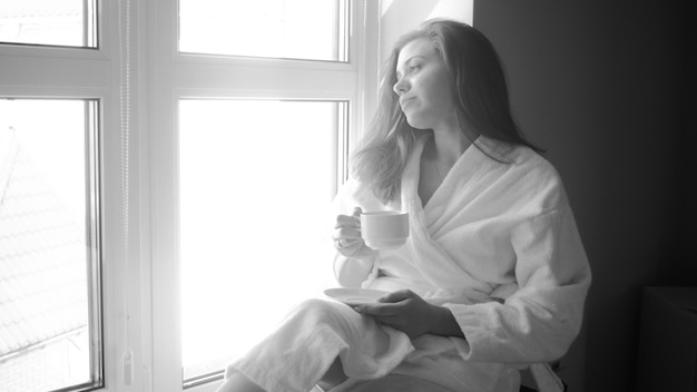 Retrato en blanco y negro de una mujer joven en bata de baño sentada en la ventana grande y tomando café