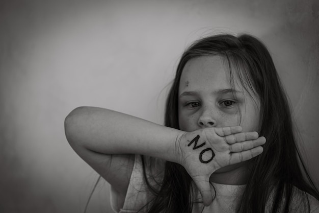 Retrato en blanco y negro de una niña magullada asustada levantando la mano para cubrir la boca mostrando la inscripción no en la palma abierta