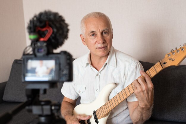 Retrato de un blogger senior tocando la guitarra desde su estudio de grabación en casa. Concepto de aprendizaje en línea.