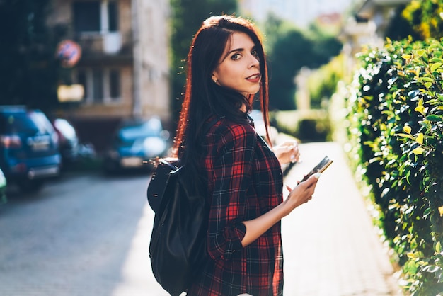 retrato de una bloguera encantadora que sostiene un teléfono inteligente moderno y lleva una mochila negra
