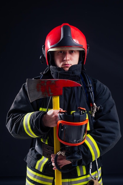 Retrato bombero fuerte en uniforme ignífugo sosteniendo un hacha en sus manos estudio de fondo negro