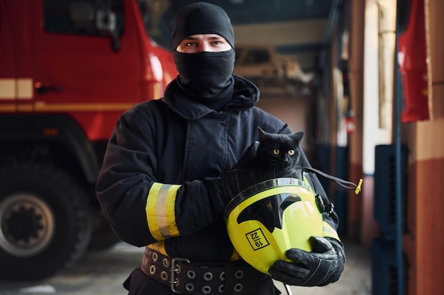 Retrato de bombero con uniforme protector que sostiene un lindo gato negro