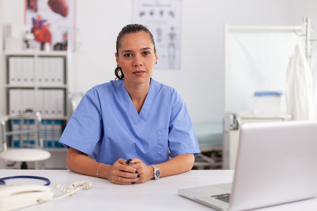 Retrato de bonita enfermera médico sonriendo al frente en la oficina del hospital vistiendo uniforme azul