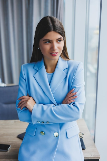 Retrato de una bonita morena con una chaqueta azul en la oficina cerca de la ventana panorámica con vista a la ciudad Mujer de negocios feliz en la oficina