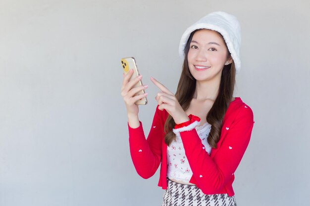 Retrato de una bonita muchacha adolescente asiática con vestido rojo y sombrero blanco sonríe felizmente con smartphone