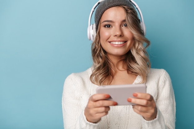 Retrato de bonita mujer alegre en gorro de lana con auriculares y teléfono móvil aislado sobre la pared azul