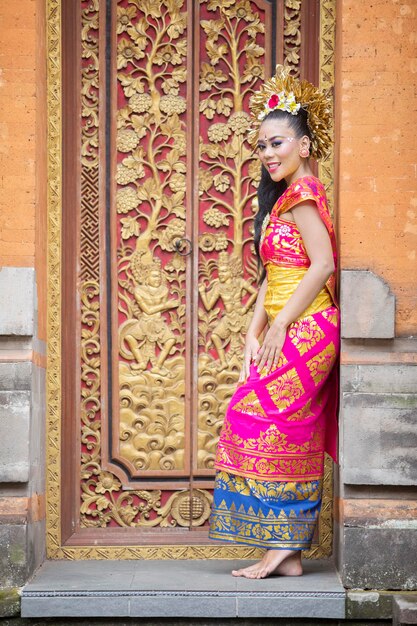 Foto retrato de una bonita mujer balinesa sonriendo a la cámara mientras lleva ropa tradicional y está de pie en la puerta de un templo