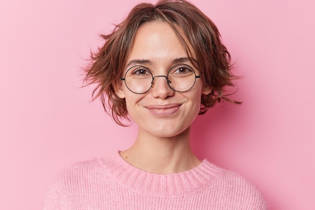 Retrato de una bonita mujer europea con sonrisas de pelo corto y oscuro que se siente satisfecha lleva anteojos redondos transparentes para la corrección de la visión y un saltador casual aislado sobre fondo rosa.