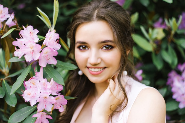 Retrato bonito de uma menina bonita que esteja cercada pelo rododendro de florescência das flores cor-de-rosa lilás na primavera no jardim. Bela pele limpa de uma jovem mulher. Maquiagem profissional