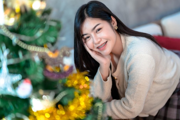 Retrato Bonito lindo sorriso positivo jovem mulher asiática decorando uma árvore de Natal em casa na sala de estar Decoração durante o Natal Natal e feriados de Ano Novo