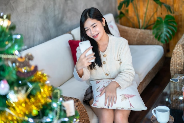 Retrato Bonito lindo sorriso positivo jovem mulher asiática usando chamada de vídeo smartphone ou Selfie em casa na sala de estar Decoração durante o Natal Natal e feriados de Ano Novo