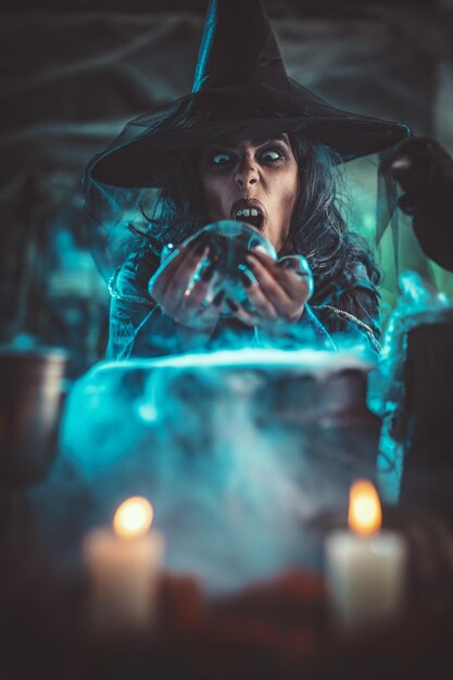 Retrato de bruja con cara terrible en un entorno ahumado espeluznante envía marcas malvadas con bola mágica sobre poción hirviendo.