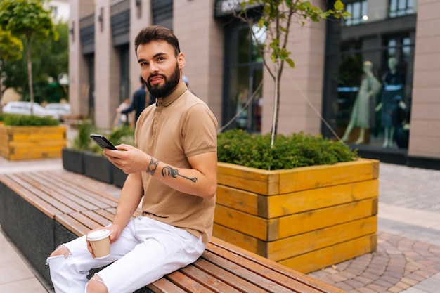 Retrato de un brutal joven barbudo sosteniendo un teléfono inteligente sentado en un banco con café para llevar mirando la cámara en la calle urbana