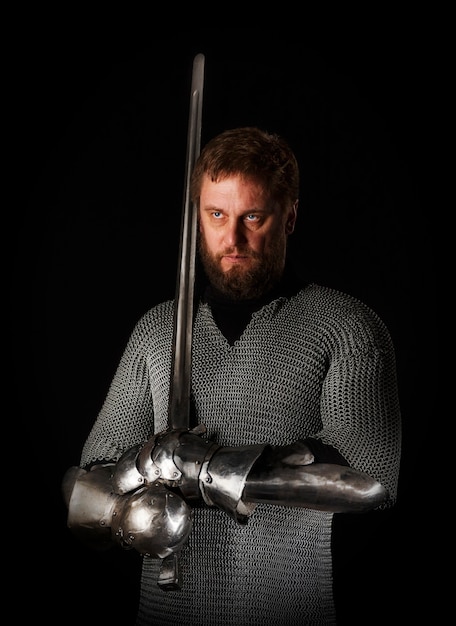 Foto retrato de un caballero medieval con barba en cota de malla con una espada de caballero en la mano y armadura, aislado en una pared oscura