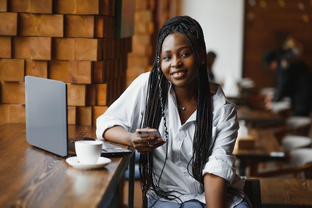 Retrato en la cabeza de una feliz y sonriente mujer afroamericana sentada en la mesa de un café mirando a la cámara emocionada mujer posando trabajando en la computadora haciendo la tarea preparando un informe en la cafetería