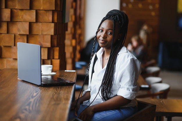 Retrato en la cabeza de una feliz y sonriente mujer afroamericana sentada en la mesa de un café mirando a la cámara emocionada mujer posando trabajando en la computadora haciendo la tarea preparando un informe en la cafetería