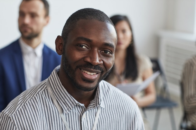 Retrato de cabeza y hombros del empresario afroamericano mirando a la cámara y sonriendo mientras está sentado en la audiencia en una conferencia de negocios o seminario, espacio de copia