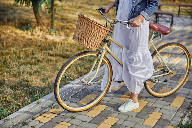 Retrato de cabeza recortada de mujer adulta en vestido blanco de pie en la carretera con bicicleta retro