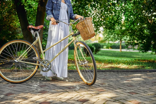 Retrato de cabeza recortada de mujer con vestido blanco y chaqueta de mezclilla caminando con bicicleta retro por el parque verde