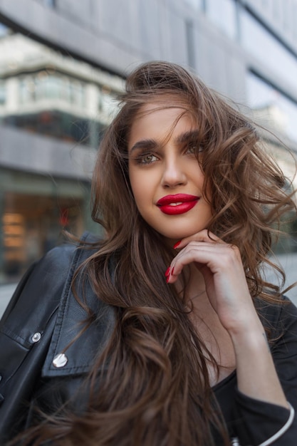 Retrato callejero femenino de belleza feliz joven elegante mujer de moda con labios rojos en ropa negra con chaqueta de cuero camina por la ciudad