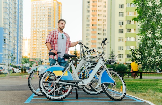 Retrato callejero de un hombre barbudo con ropa informal parado en un estacionamiento de bicicletas y dando un paseo en bicicleta mirando a la cámara con una cara seria El hombre adulto usa el alquiler de bicicletas