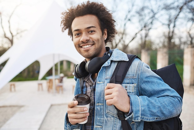 Retrato callejero de un joven de piel oscura positivo y atractivo que sostiene una mochila y un café mientras pasea por el parque con su novia en busca de un lugar tranquilo Banda después de practicar discutiendo música