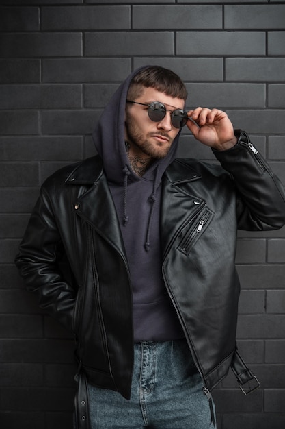 Retrato callejero de moda de hombre guapo hipster con gafas de sol elegantes y frescas en una chaqueta de cuero de moda con poses de capucha cerca de un fondo de ladrillo moderno negro