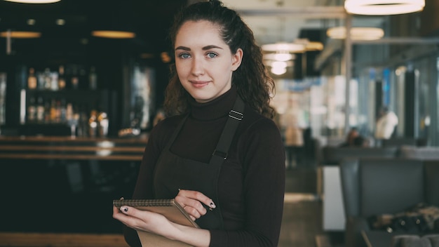 Retrato de una camarera joven alegre y hermosa de pie manteniendo el menú y sonriendo sinceramente en un café astuto