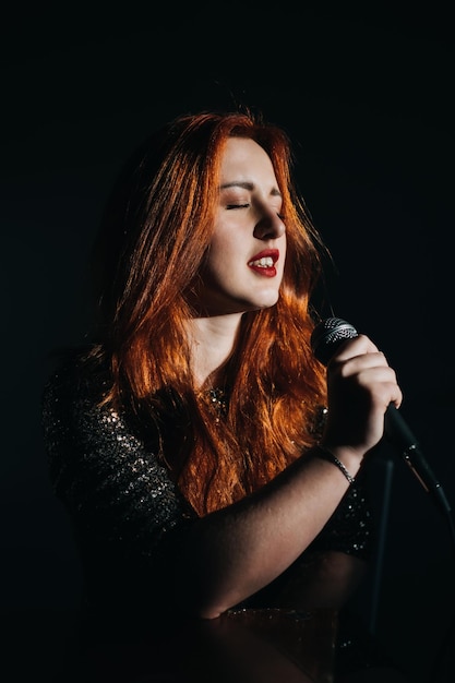 Retrato de una cantante pelirroja con un vestido de noche brillante sosteniendo un micrófono en la noche oscura