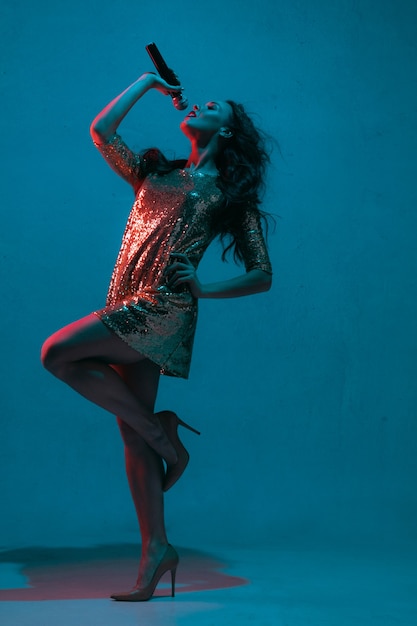 Retrato de cantante de raza caucásica aislado sobre fondo azul de estudio en luz de neón. Modelo de mujer hermosa en vestido brillante con micrófono. Concepto de emociones humanas, expresión facial, publicidad, música, arte.